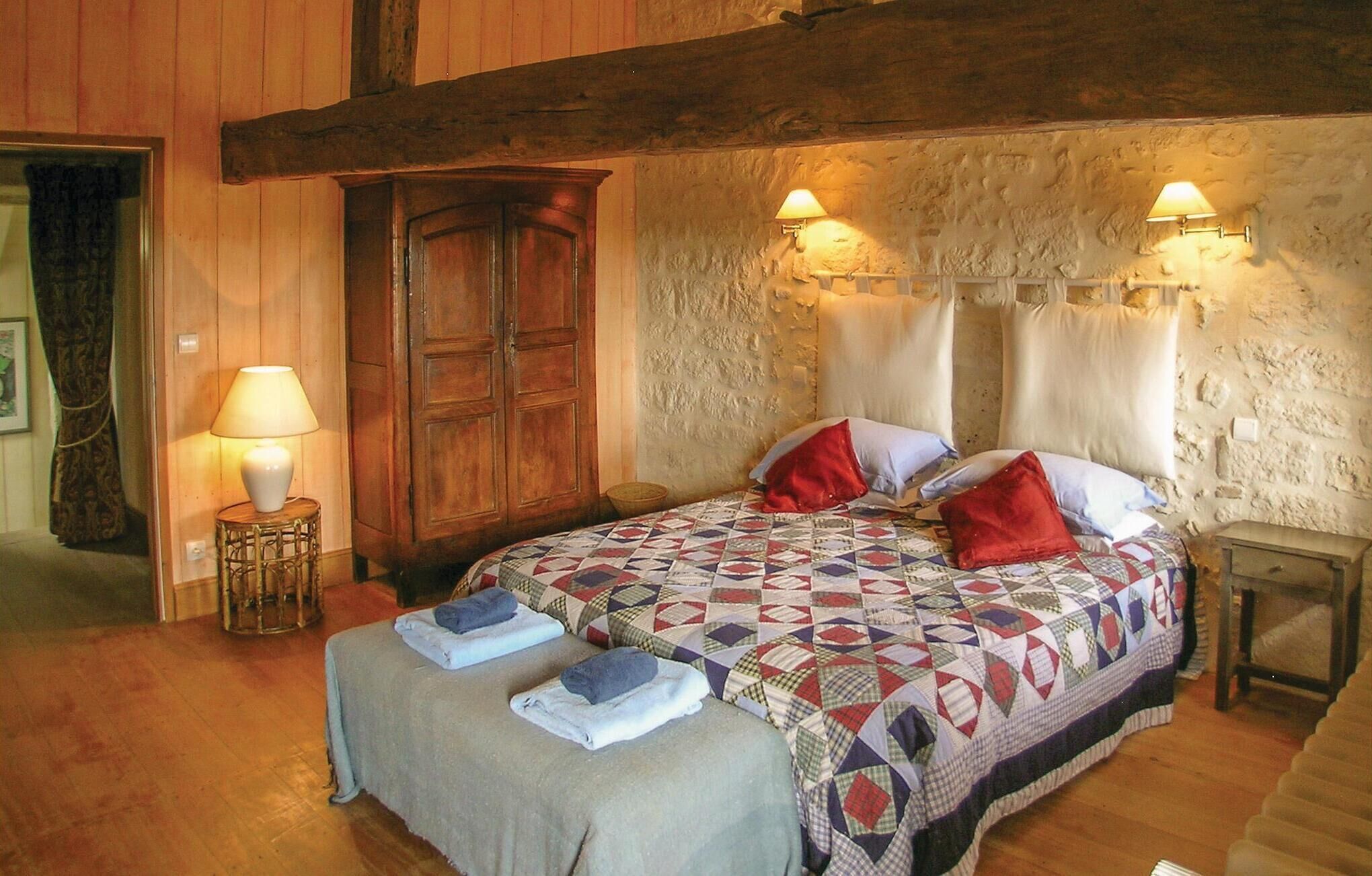 A cozy bedroom with a wooden bed, a quilt, two bedside lamps, and a stone wall.