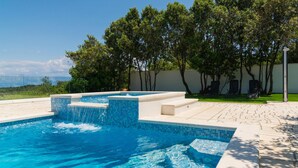 A swimming pool with clear blue water, surrounded by a tiled deck and a grassy area with trees.