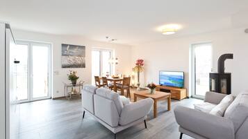 Ein modernes Wohnzimmer mit einer Couch, einem Couchtisch und einem Fernseher. Es gibt einen Essbereich mit Stühlen und einem Tisch, sowie eine Küchenzeile mit einem Herd.