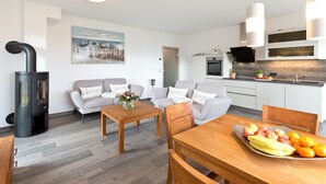 A modern living room with a sofa, a dining table with fruit, a kitchen with a stove and cabinets, and a painting on the wall. - Great Ocean - Kells apartments (Klink)