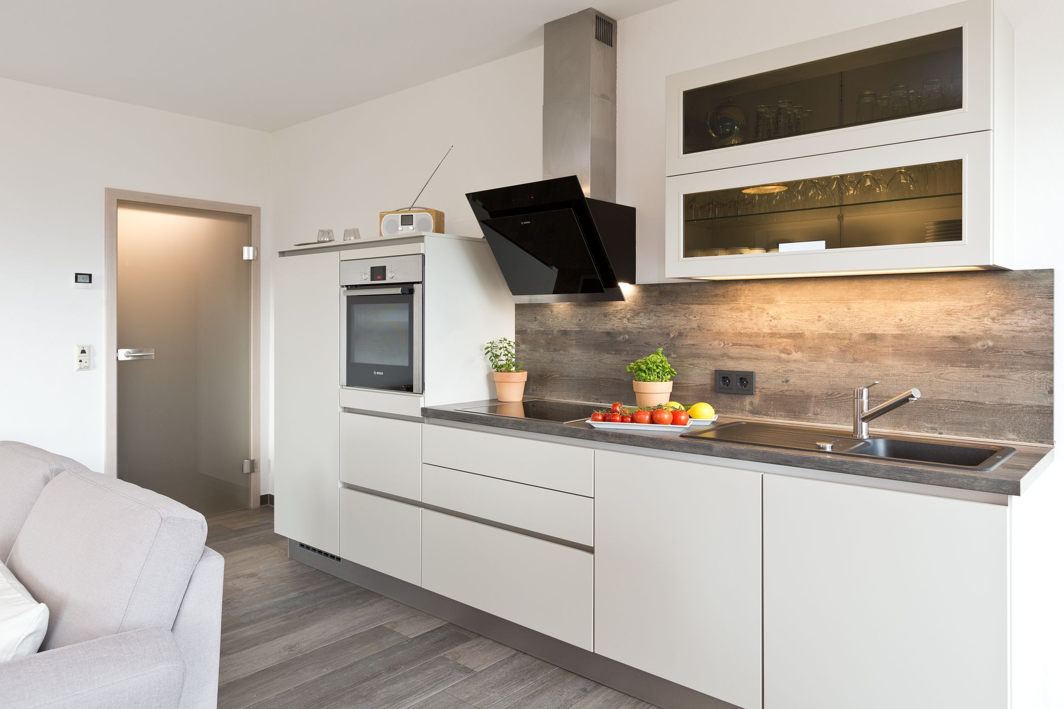 Une cuisine moderne avec des meubles blancs, un four encastré, un four à micro-ondes, une hotte noire et un pare-feu en bois.