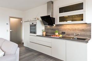 A modern kitchen with white cabinetry, a built-in oven, a microwave, a black range hood, and a wooden backsplash. - Great Ocean - Kells apartments (Klink)