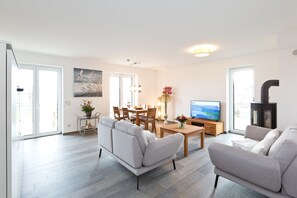 A modern living room with a sofa, a coffee table, and a TV. There is a dining area with chairs and a table, and a kitchen area with a stove. - Great Ocean - Kells apartments (Klink)