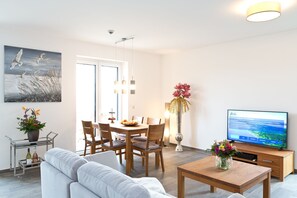 A modern living room with a sofa, dining table, chairs, a TV, and a vase of flowers. - Great Ocean - Kells apartments (Klink)