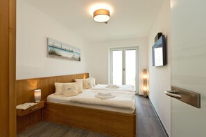 A hotel room with a wooden headboard, a painting of a beach scene, a flat-screen TV mounted on the wall, and a bedside table with a lamp. - Great Ocean - Kells apartments (Klink)