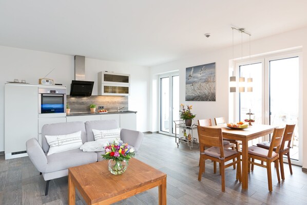 A modern living room with a sofa, coffee table, and dining table with chairs. There is a kitchen area with a stove, oven, and microwave. A TV is mounted above the stove.
