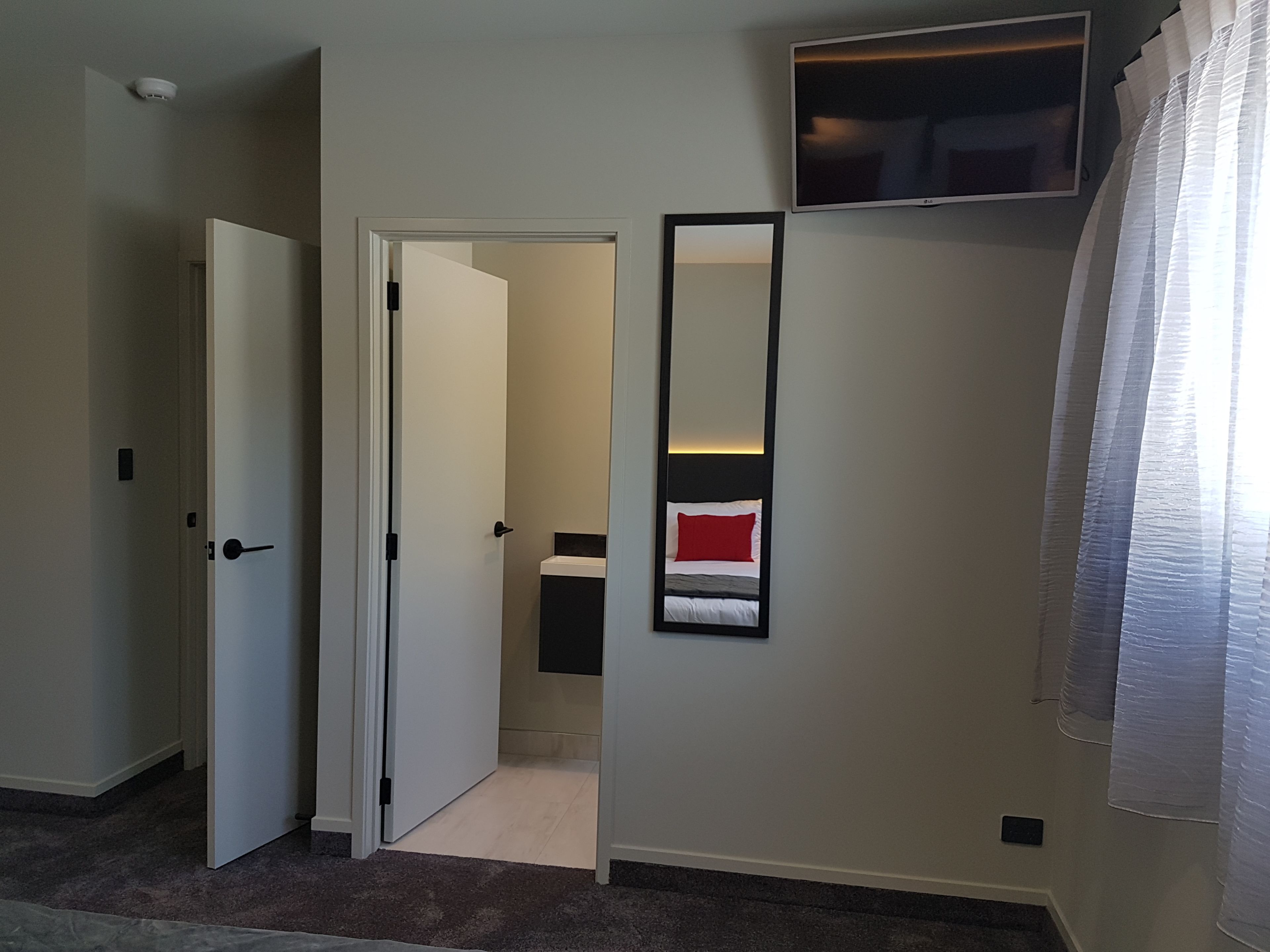 A hotel room with a closed door, a mirror on the wall, a TV mounted above the mirror, and a window with sheer curtains.