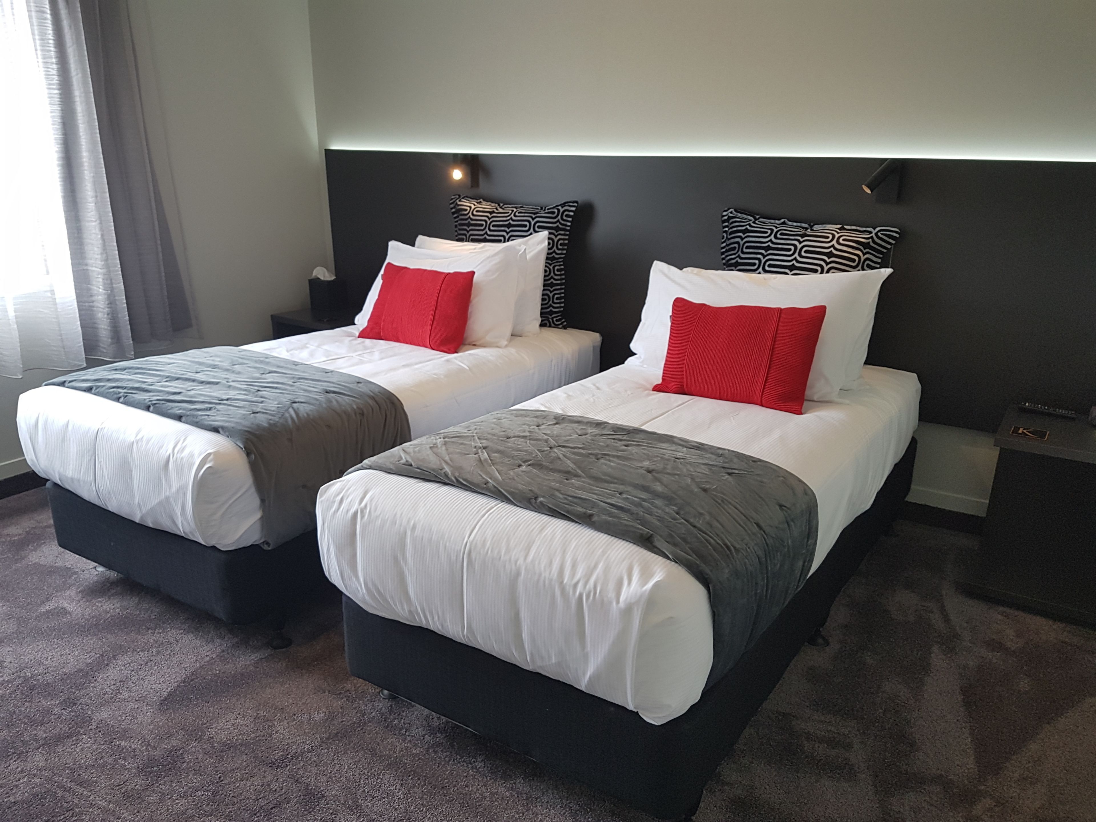 Two double beds with grey and white bedding, red pillows, and black headboards in a hotel room.