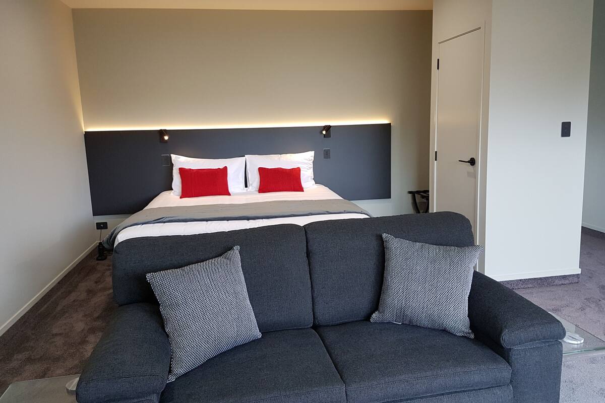 A modern hotel room with a large bed, grey sofa, and a white door.