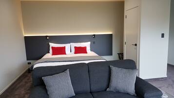 A modern hotel room with a large bed, grey sofa, and a white door.