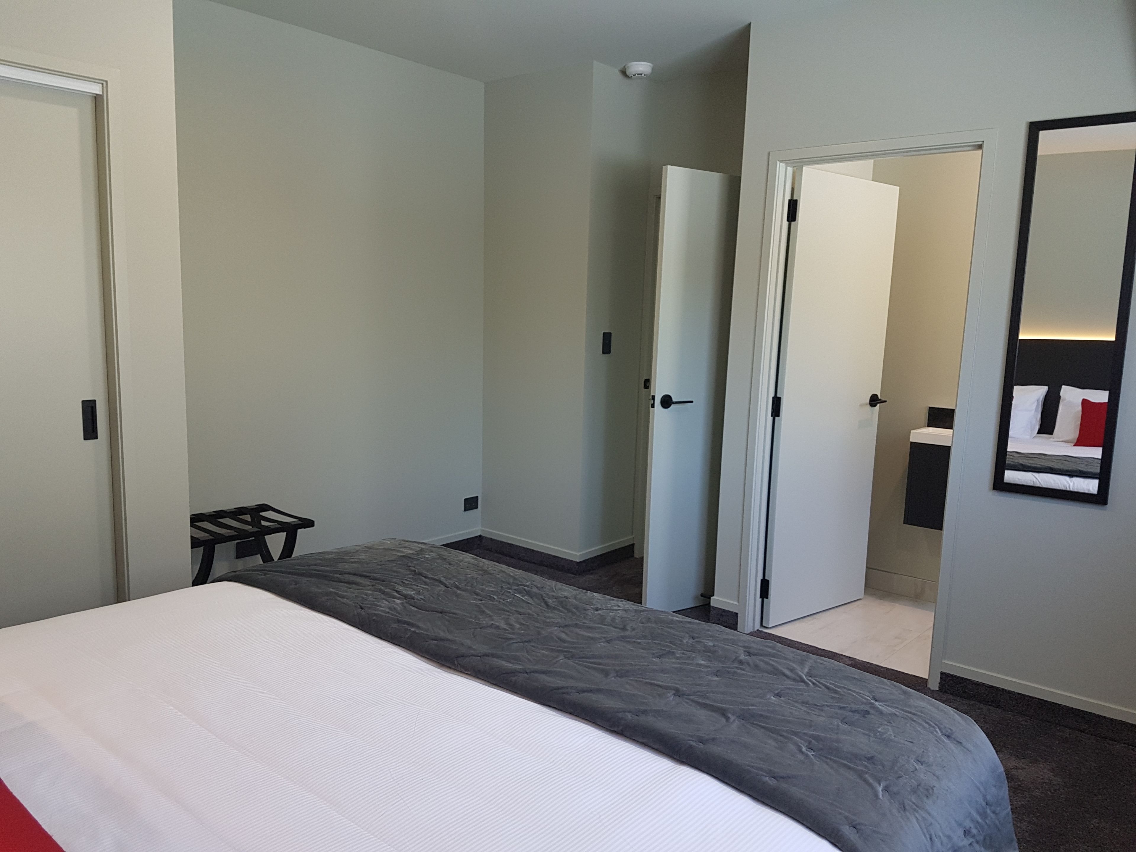 A hotel room with a bed, a small table, a mirror, and a door leading to a bathroom.