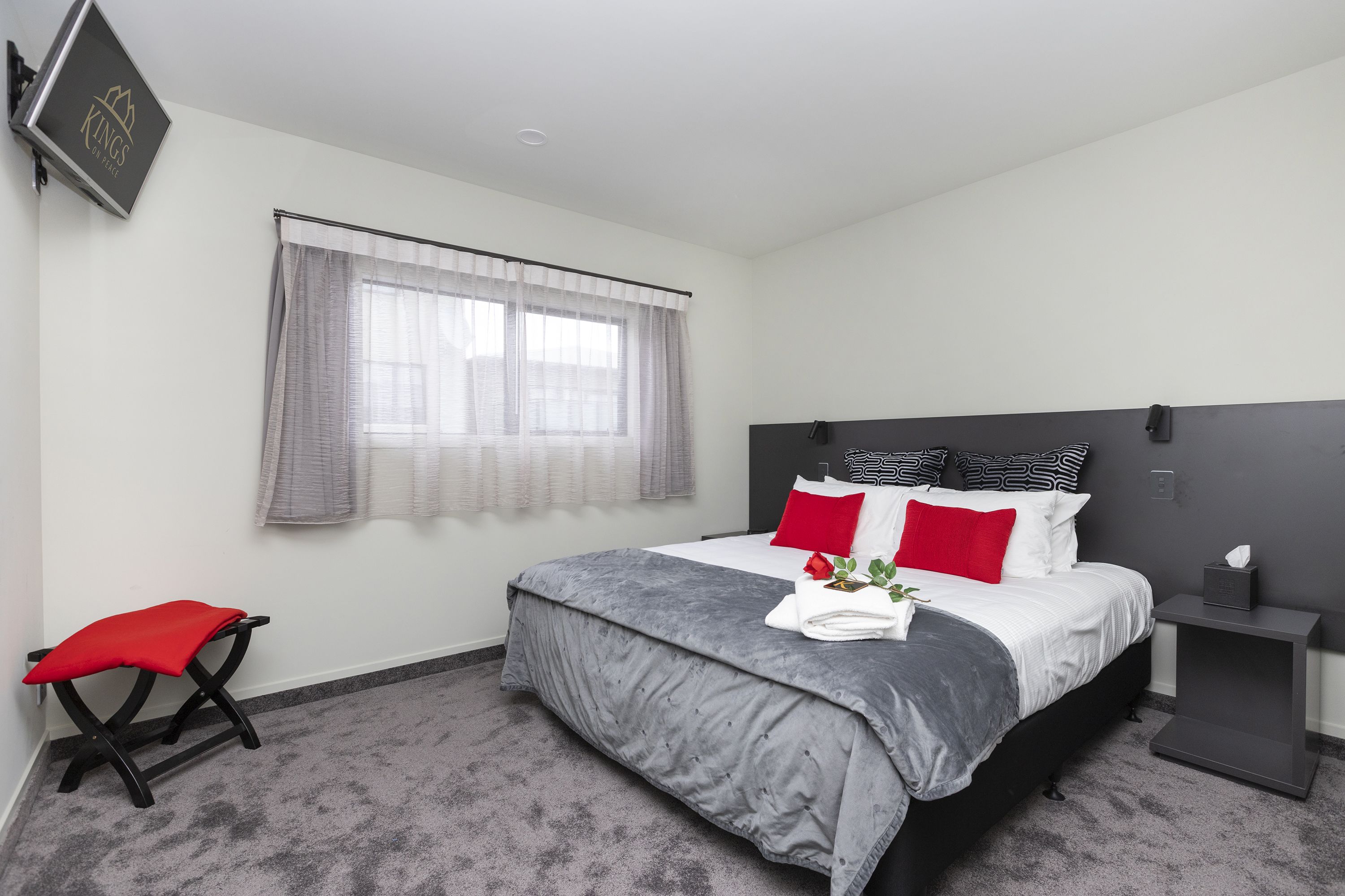 A modern bedroom with a bed, a nightstand, a red chair, and a window with sheer curtains.