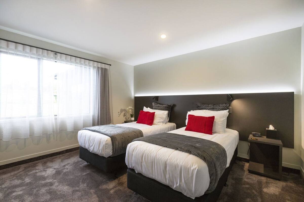 A hotel room with two beds, a nightstand, a lamp, and a window with curtains.