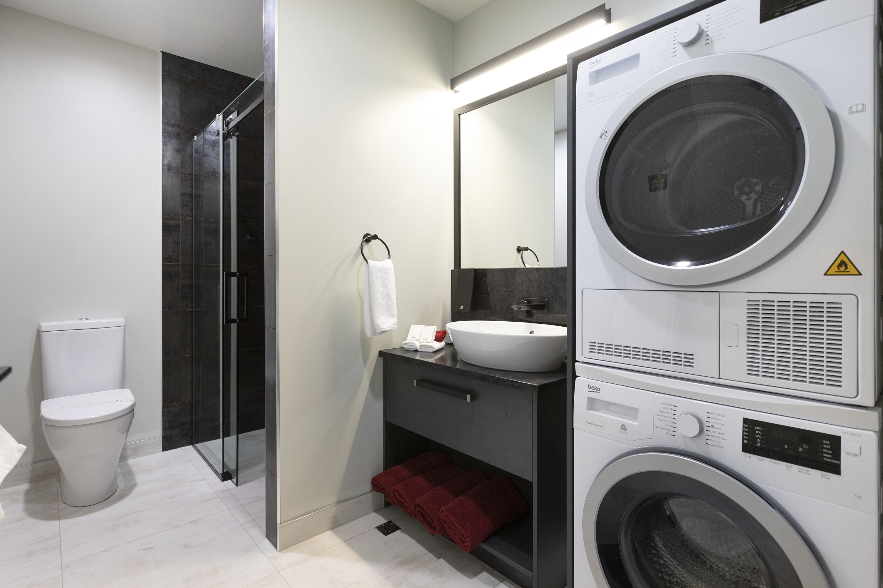 A modern bathroom with a toilet, a glass shower enclosure, a sink, and a stacked washer and dryer.