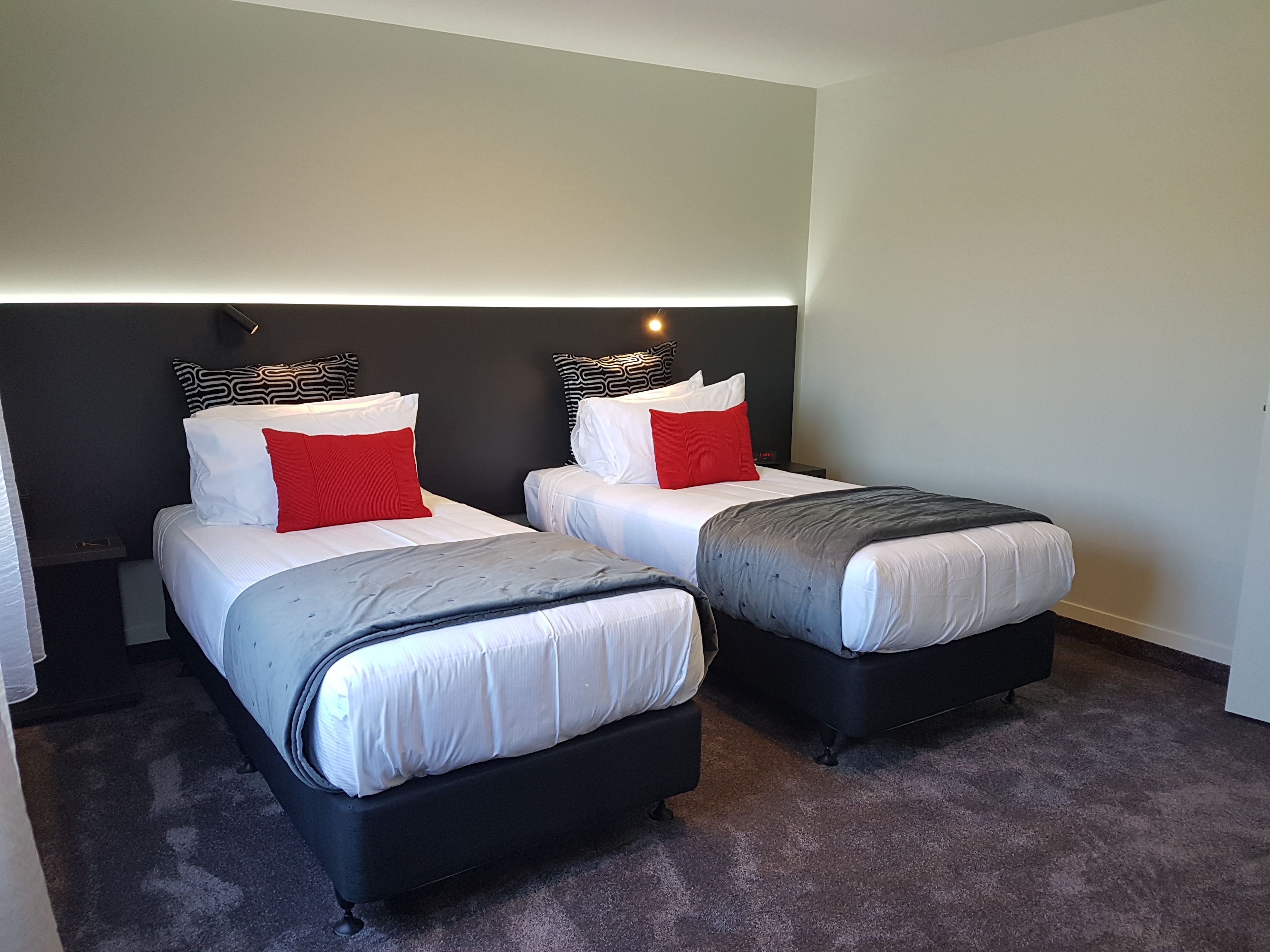 A hotel room with two beds, each with white bedding and red pillows, a dark headboard, and a carpeted floor.