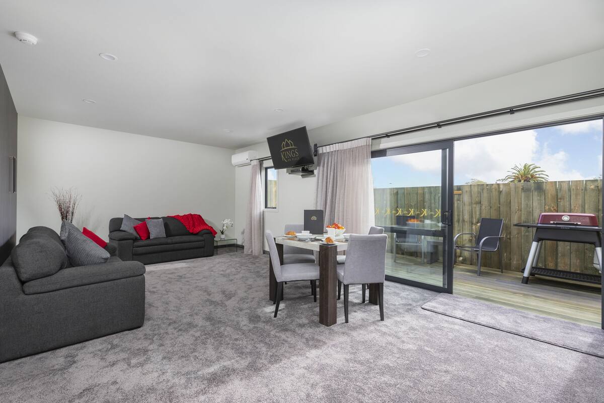 A modern living room with a grey sofa, a dining table with chairs, a flat-screen TV, and a sliding glass door leading to an outdoor patio.