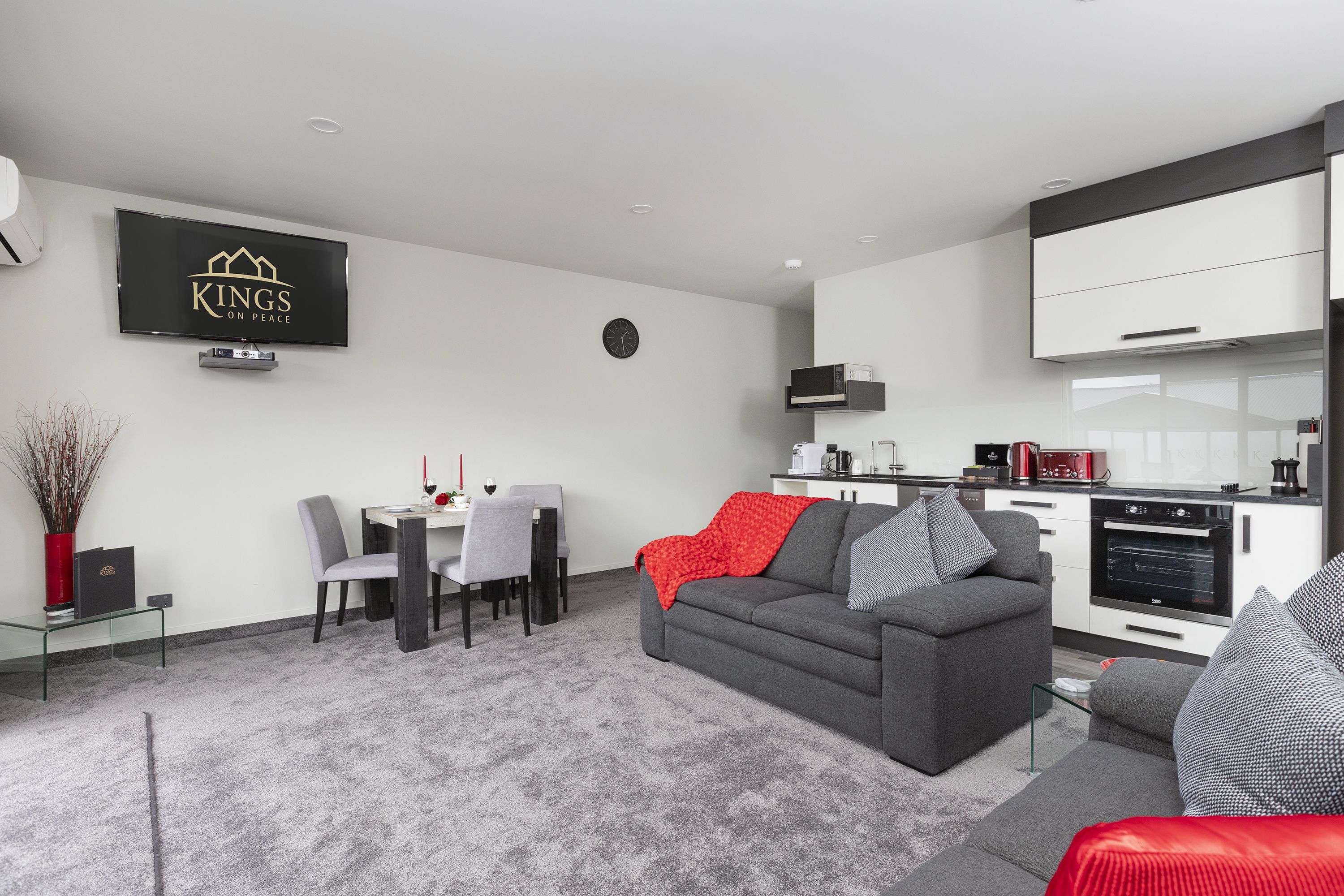 A modern living room with a grey sofa, a dining area with a table and chairs, and a kitchen with white cabinets and appliances.
