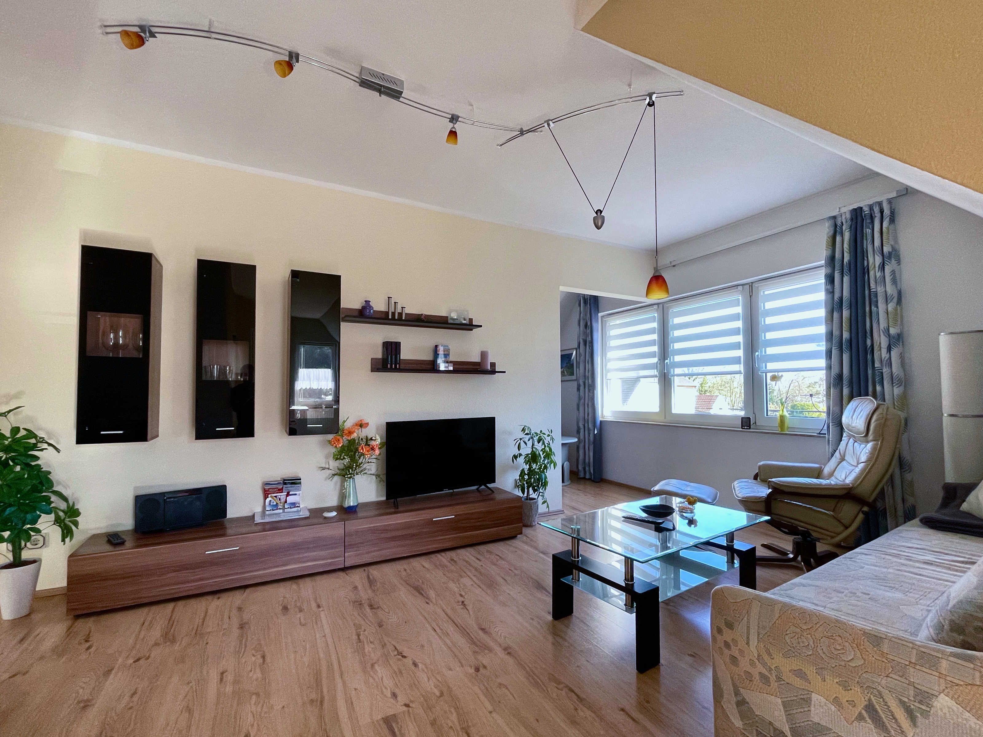 Una sala moderna con televisor de pantalla plana, mueble de entretenimiento de madera, mesa de centro de vidrio, sofá de cuero y un ventanal con persianas.