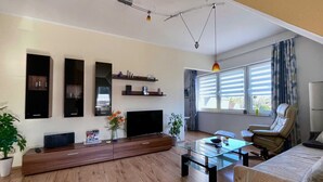 Ein modernes Wohnzimmer mit Flachbildfernseher, Holz-Entertainment-Möbel, Glastisch, Ledersofa und einem großen Fenster mit Jalousien.