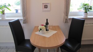 Un pequeño comedor con una mesa de madera, dos sillas negras, una botella de vino y un florero con flores.
