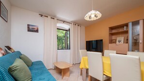 Ein gemütliches Wohnzimmer mit einem blauen Sofa, einem kleinen Holztisch und einem Esstisch mit gelbem Tischleineneindeckung und Stühlen. Ein Fernseher ist an der Wand angebracht, und es gibt ein Bücherregal mit Dekorationselementen.
