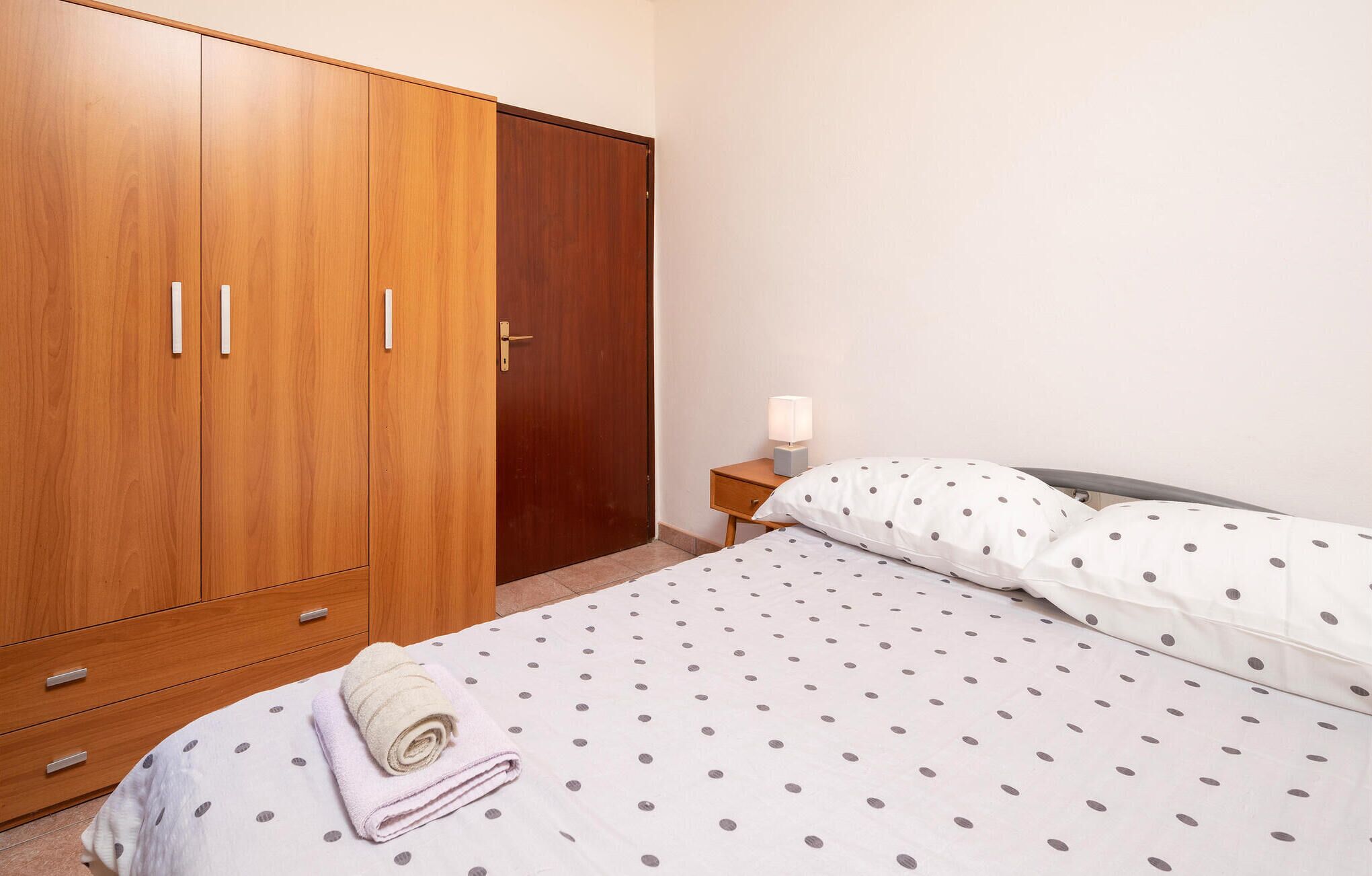 Un dormitorio con un armario de madera, una mesita de noche con lámpara, una cama con ropa de cama de lunares y una toalla enrollada en el suelo.