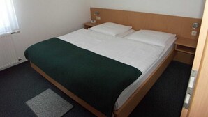 En enkel seng med trægavle, hvidt sengetøj og et grønt plaide. Der er et lille natbord med en lampe, et indrammet billede på væggen og en radiator i hjørnet.