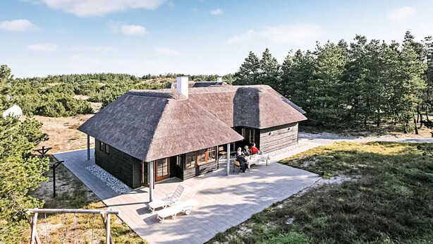 A rustic cabin with a thatched roof, a patio with outdoor seating, and a view of the surrounding forest.