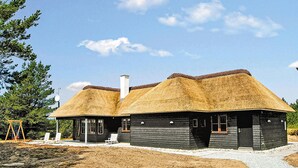 A rustic cabin with a thatched roof, a chimney, and a small porch with white chairs.