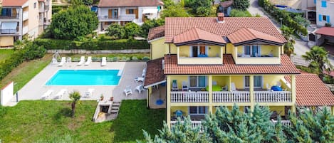A two-story house with a swimming pool, surrounded by greenery and other houses.