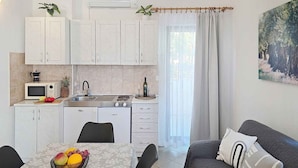 A compact kitchen with a microwave, stove, sink, and cabinets. There is a dining table with fruit and a couch with pillows.