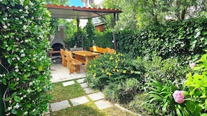 A garden patio with a stone table and chairs, a brick oven, and a pergola.