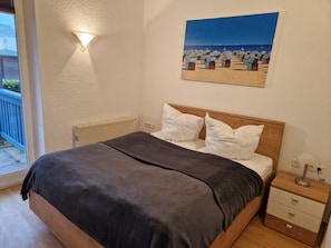 A bedroom with a bed, a nightstand, a wall-mounted lamp, and a beach scene painting. - (22) 2-room apartment - apartment house Sonnenresidenz I (Kühlungsborn)