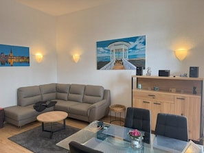 A modern living room with a sectional sofa, a glass coffee table, and a dining area with a glass table. There are two paintings on the wall, a wooden cabinet, and a small stool. - (22) 2-room apartment - apartment house Sonnenresidenz I (Kühlungsborn)