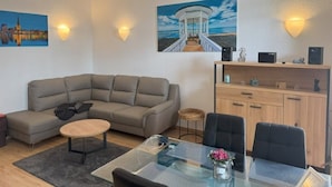Ein modernes Wohnzimmer mit einem Ecksofa, einem Glastisch und einem Essbereich mit einem Glastisch. An der Wand befinden sich zwei Gemälde, eine Holzkommode und ein kleiner Hocker.