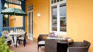 Eine Terrasse im Freien mit Korbmöbeln, einem Tisch mit Blumenvase und einem blauen Sonnenschirm.