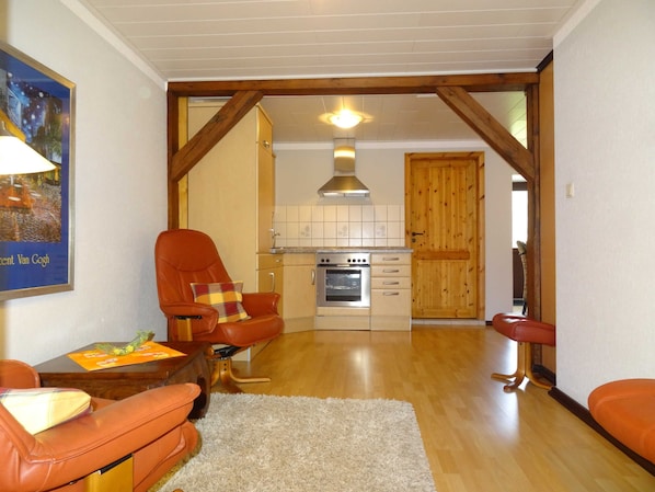 A living room with a sofa, a chair, a small table, and a kitchen area with a stove and cabinets.