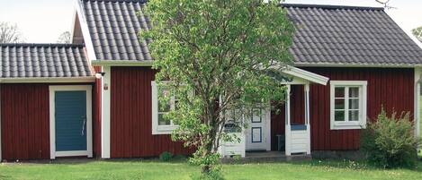 Une maison à deux étages, avec une façade rouge, une antenne parabolique et un petit arbre dans le jardin.