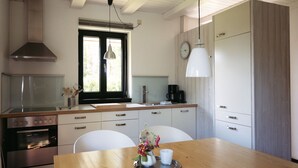 Et moderne køkken med et træbord, hvide stole, en stålfarvet ovn og et vindue med udsigt over grønne områder.