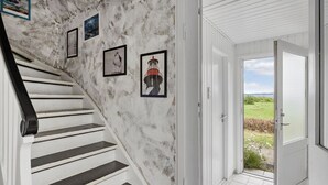 Un escalier aux murs blancs et aux marches noires mène à une porte qui donne sur un paysage verdoyant.