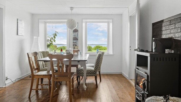Un coin repas avec une table et des chaises en bois, deux fenêtres donnant sur un paysage verdoyant et une cheminée sur le côté droit.