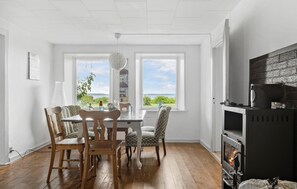 A dining area with a wooden table and chairs, two windows with a view of greenery, and a fireplace on the right. - 3 bedroom amazing apartment in Broager (Broager)