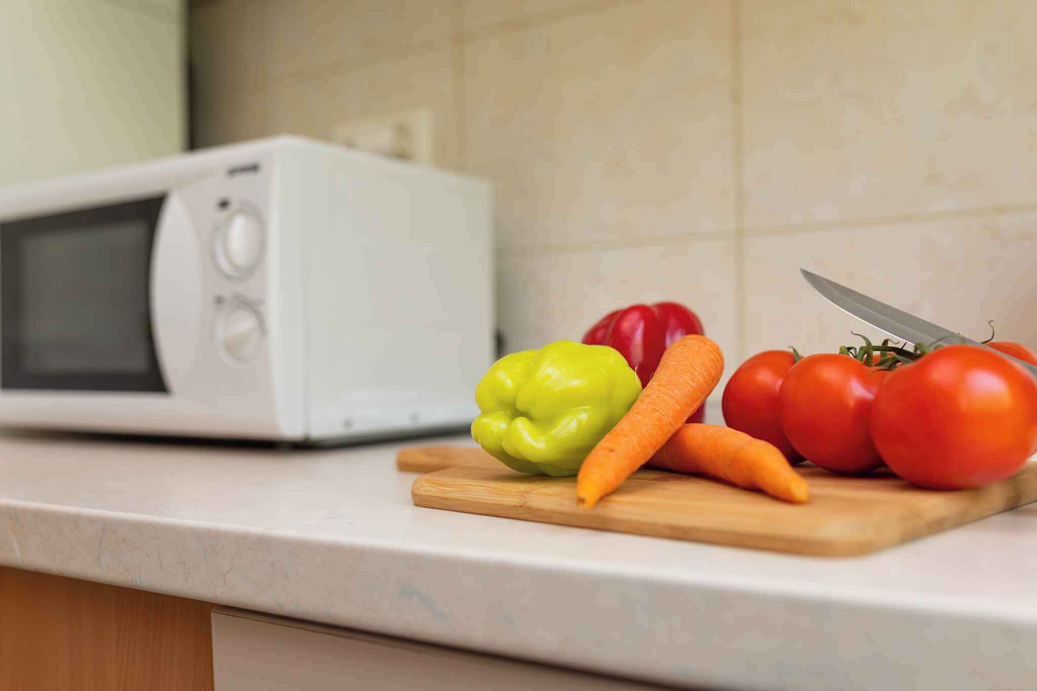 Una encimera de cocina con un microondas, una tabla de cortar con verduras y un cuchillo.