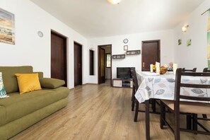 Een woonkamer met een groene bank, houten vloeren, een eettafel met stoelen, een televisie en een klok aan de muur.