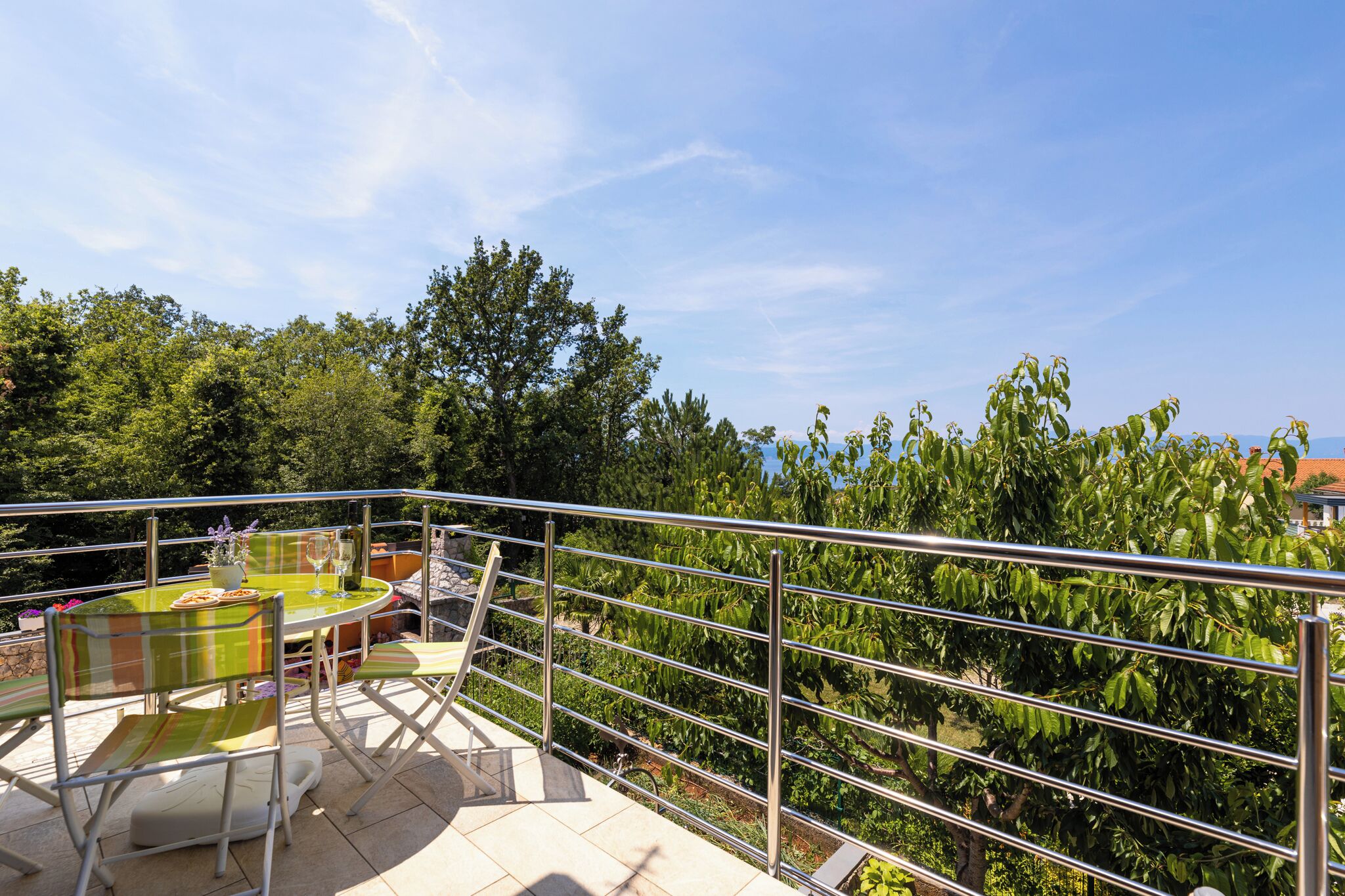 A balcony with a table and chairs, surrounded by metal railings, overlooking a lush garden and trees.
