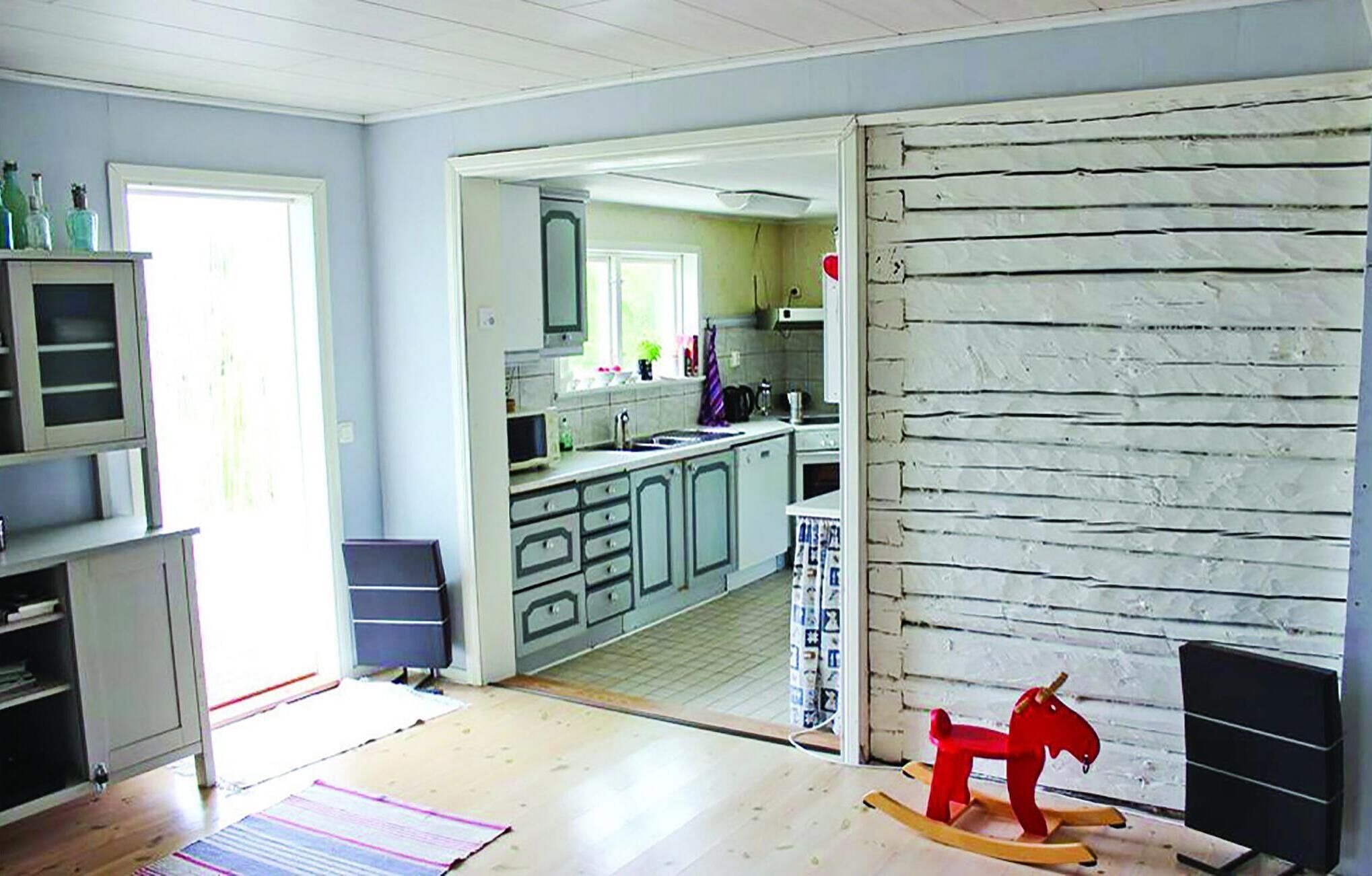 Une pièce avec un plancher en bois, un cheval à bascule, un coin cuisine avec des placards et une fenêtre, et une porte donnant sur une autre pièce.