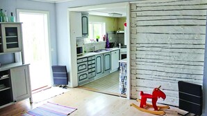 Una habitación con piso de madera, un caballo balancín, una zona de cocina con armarios y una ventana, y una puerta que da a otra habitación.