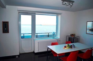 A room with a large window overlooking the sea, a white table with red chairs, and a small kitchenette. - Sino Suite, 2. OG, balcony seafront, - Dünenblick Apartments (Helgoland)