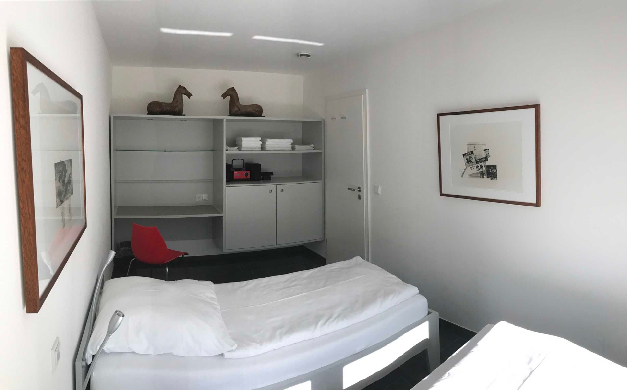 Et lille, pænt ordnet værelse med en seng, en rød stol, et garderobeskab og et billede i ramme på væggen.