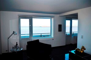 A room with a large window offering a view of the sea, a radiator, a lamp, and a small table with a bottle. - Sino Suite, 2. OG, balcony seafront, - Dünenblick Apartments (Helgoland)